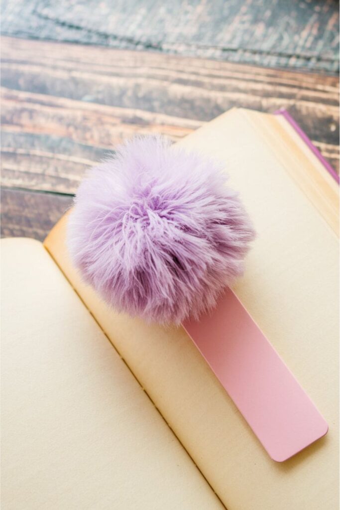 A fluffy pom-pom bookmark made from soft yarn, attached to a long ribbon or thread, bringing a cozy and playful touch to a book