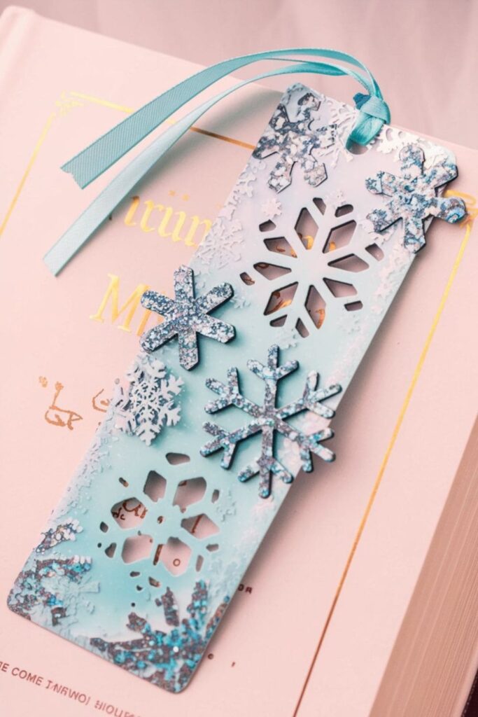 A winter-themed bookmark in icy blue and silver shades, decorated with intricate snowflake cutouts and glitter to create a frosty, magical effect