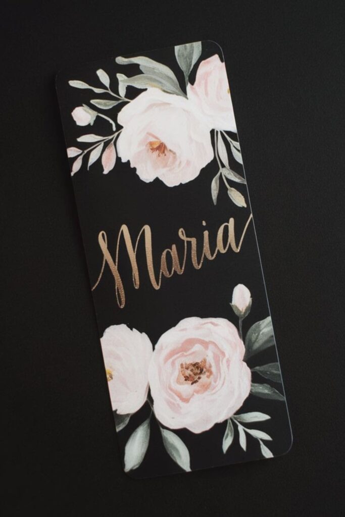 Personalized Floral Bookmarks A sleek floral bookmark with the name _Maria_ in gold calligraphy on a deep matte black background, adorned with delicate blush pink and white flowers